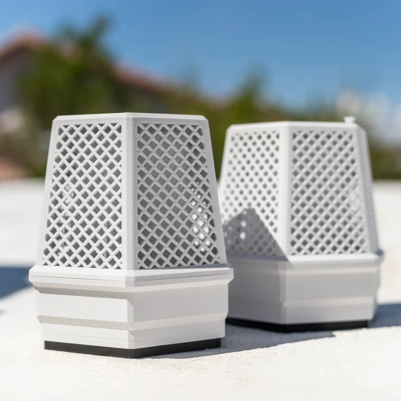 Two white BuzzBox mosquito control units with lattice design, placed outdoors on a bright surface with a blurred house and trees in the background under a blue sky.
