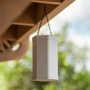White ScoutStop mosquito control unit hanging from a chain under a roof eave, with a blurred green outdoor background.