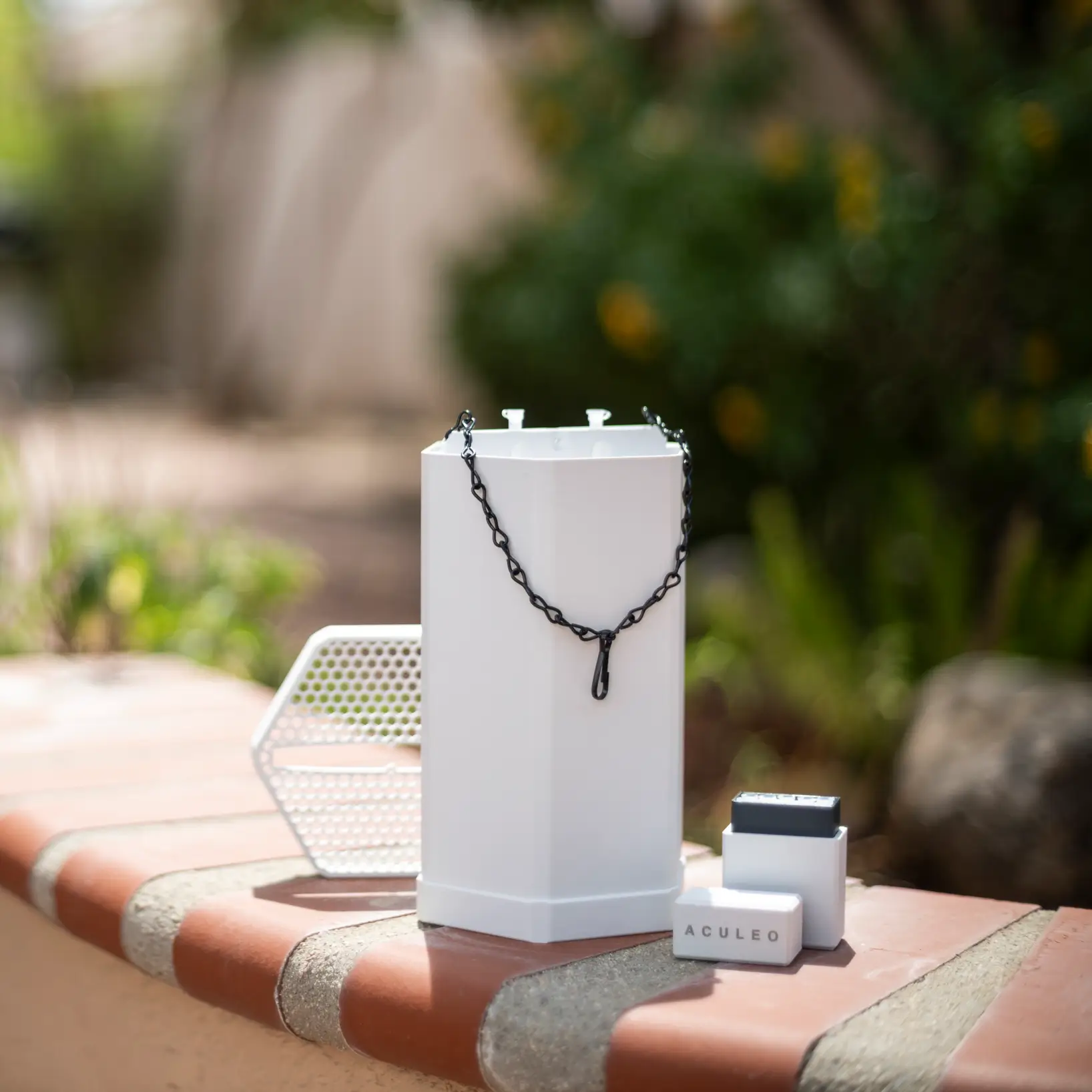 White electronic device with a chain, labeled components, and a honeycomb-patterned accessory placed on a brick ledge.